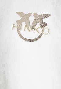 Valkoinen ribbikangas, jossa on kultaisella brodeerattu logo, jossa esiintyy kaksi lintua ja brändin nimi "PINKO" käsinkirjoitettuna, ympyräkehyksen yksityiskohdalla.