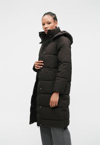 Melns polsterēts garais mētelis ar kapuci, ar rāvējslēdzēju, spiedpogu aizdari un sānu kabatām. Audumam ir gluda, matēta virsma.