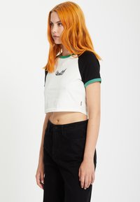 Gekürztes T-Shirt mit einem weißen Körper und schwarzen Ärmeln, grünem Kragen und Flügelgrafik auf der Vorderseite. Kombiniert mit schwarzen hochgeschnittenen Hosen.