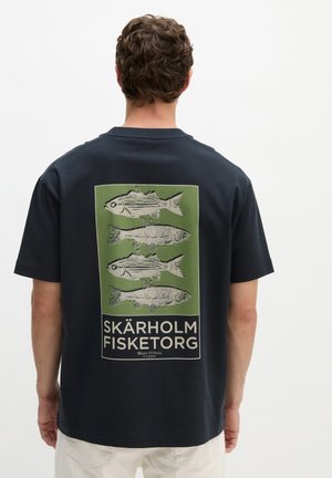 Homme portant une chemise foncée avec quatre poissons illustrés et le texte « Skärholm Fisketorg » sur un carré vert dans le dos.