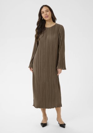 Mujer con cabello largo y oscuro que lleva un vestido midi marrón plisado de manga larga y zapatos planos negros puntiagudos sobre un fondo liso.