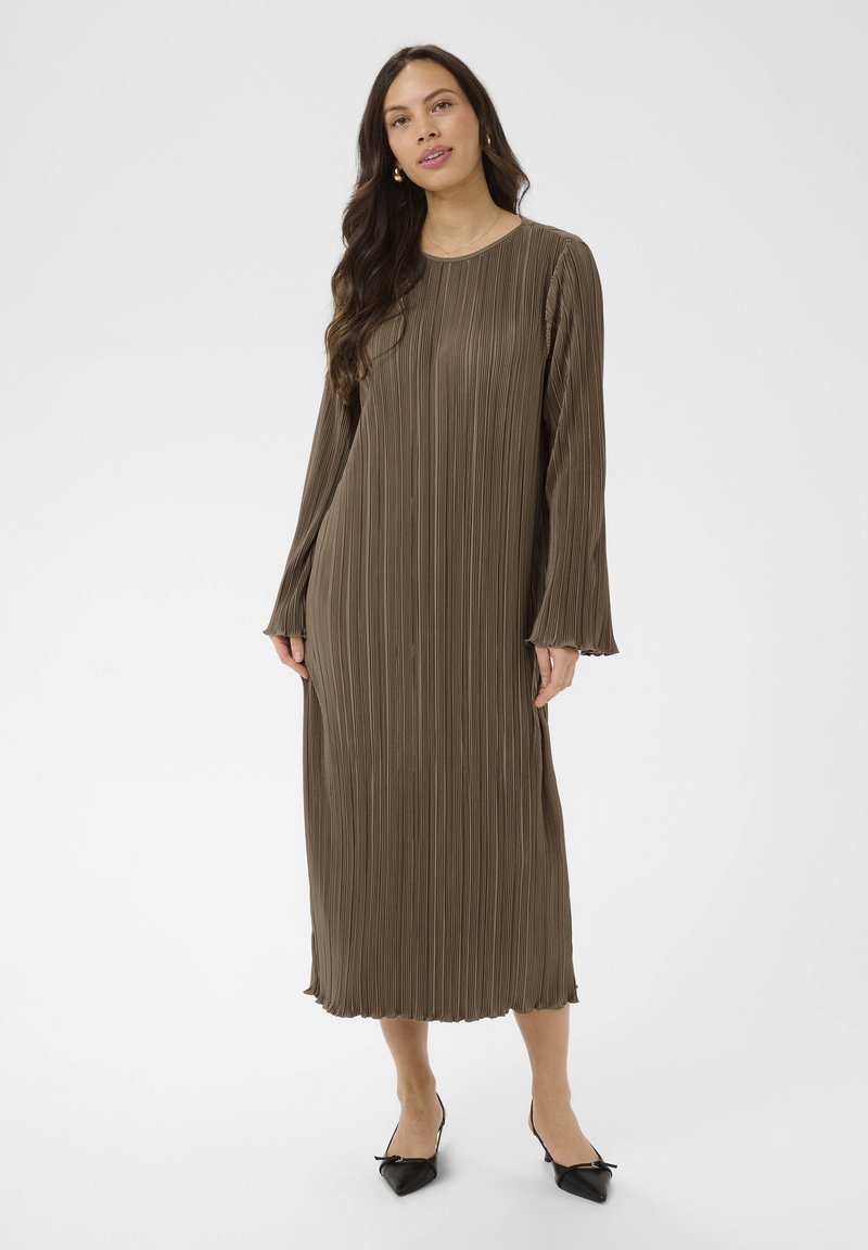 Femme aux longs cheveux foncés portant une robe midi plissée marron à manches longues et des ballerines noires pointues sur un fond uni.