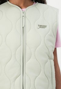 Gilet matelassé vert clair avec un col haut, doté d'une fermeture éclair à l'avant et d'un logo "tommy jeans" en relief de couleur vert foncé.