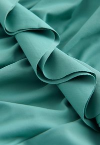 Türkiz színű szövet sima textúrával, lágy, hullámzó hajtásokkal és ívelt szélekkel, amelyek gyengéd fodrozódásokat hoznak létre.
