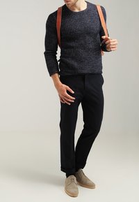 Granatowy sweter z teksturowanego dzianiny, ciemne spodnie, jasnobrązowe brogue. Brązowe skórzane paski noszone na ramionach, ręce na biodrach.