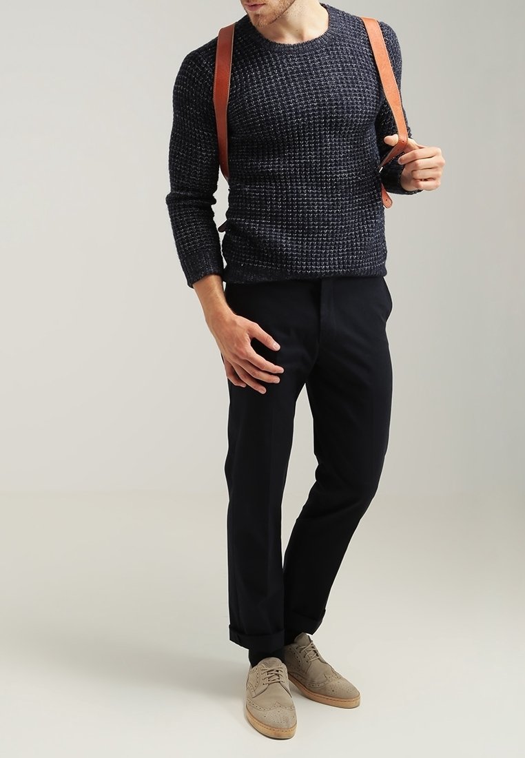 Granatowy sweter z teksturowanego dzianiny, ciemne spodnie, jasnobrązowe brogue. Brązowe skórzane paski noszone na ramionach, ręce na biodrach.