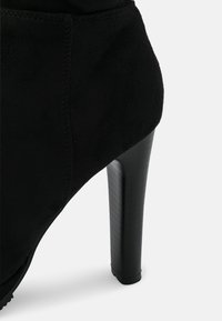 Botín de tacón alto negro con textura de ante, punta puntiaguda y tacón stiletto negro brillante. Detalles que incluyen costuras visibles.