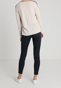 Helle beige Strickjacke mit braunen Schulterakzenten, kombiniert mit dunkelblauen Skinny-Jeans und weißen Sneakers, die einen lässigen Schnitt und eine weiche Textur zeigen.
