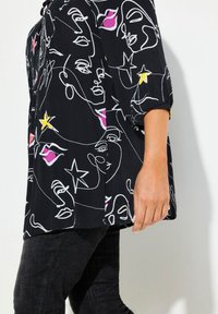 Schwarze Bluse mit einem weißen Linienkunst-Design von Gesichtern und Sternen, akzentuiert mit Pink und Gelb. Lange Ärmel mit gerafften Manschetten.