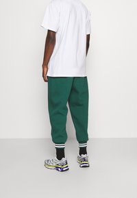 Personne portant un t-shirt blanc, un pantalon de jogging vert foncé avec des poignets rayés, des chaussettes noires et des chaussures de course Salomon grises et jaunes.