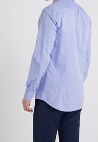 Man in een lichtblauwe lange mouwen overhemd met knopen en donkerblauwe broek, gezien van achteren tegen een effen achtergrond.