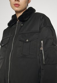 Femme portant une veste noire avec un col en fourrure, une fermeture éclair frontale, des poches à rabat et une poche zippée sur la manche gauche.