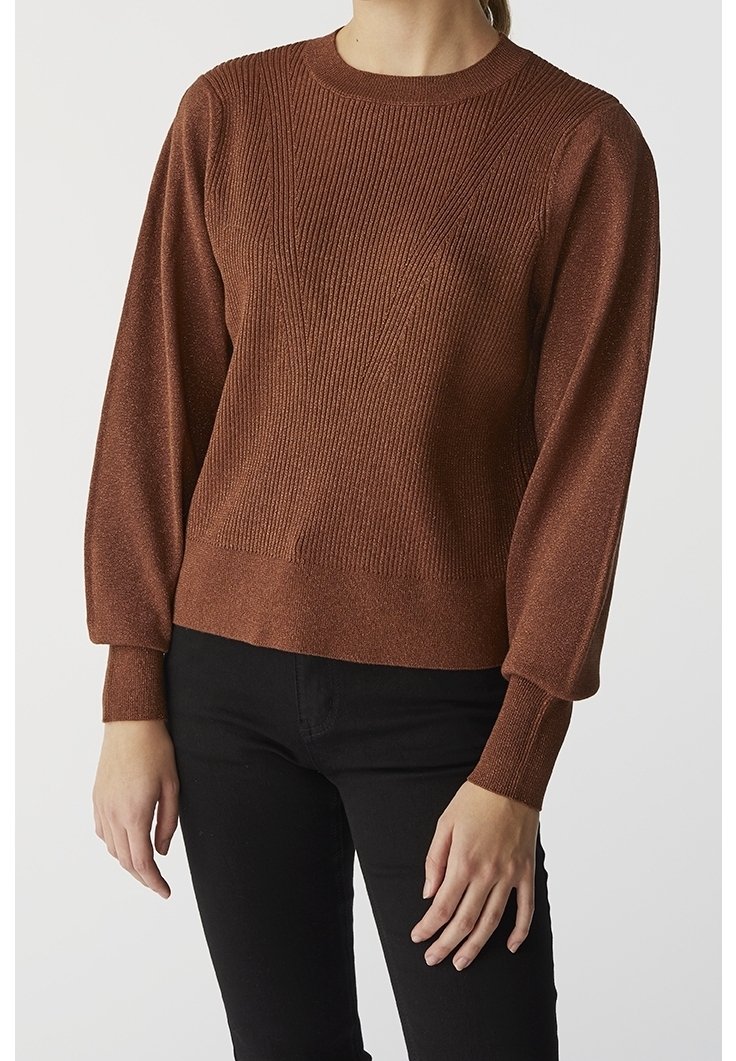 Femme portant un pull marron à manches longues côtelé avec des manches bouffantes et un pantalon noir, debout devant un fond uni.