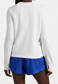 Bílá košile s dlouhým rukávem a hladkým povrchem, s kolečkovým výstřihem a bočními rozparky. Spárována s modrými sportovními šortkami s elastickým pasem.