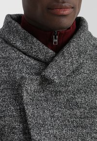 Grijze wollen jas met een grote kraag, met een subtiel structuurpatroon. Gelaagd over een donkerrode sweater met rits. 