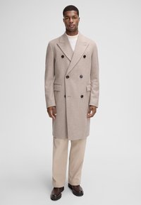 Cappotto beige in lana, doppiopetto, con grandi bottoni neri, tasche laterali e vestibilità su misura. Abbinato a pantaloni beige a coste e stivali marroni.