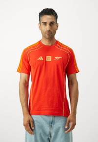 Rood T-shirt met korte mouwen en gele accenten, met het Adidas-logo. Het heeft een ronde hals en drie strepen op de schouders, gemaakt van zacht materiaal.