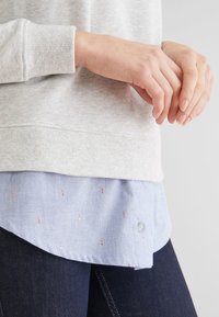 Sweat-shirt en coton gris clair avec poignets côtelés porté sur une chemise bleue à boutons avec des motifs roses subtils et un ourlet arrondi. Jean foncé visible.