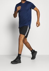 Muž jde v černých šortkách Adidas s bílými pruhy, námořnickém modrém tričku Adidas, černých ponožkách Adidas a černých běžeckých botách.