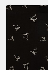Tissu noir avec un motif répétitif de nœuds blancs, présentant une texture lisse et des coutures régulières, idéal pour une tenue décontractée.