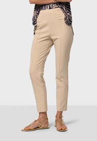 Pantalons beiges ajustés avec des coutures latérales, une texture lisse et une coupe slim, associés à des sandales plates dorées. Design léger et polyvalent.