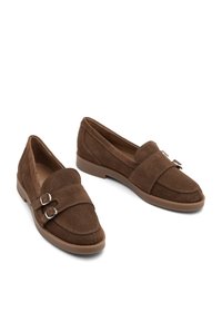 Un paio di mocassini in suede marrone con doppie fibbie argentate su una scarpa, suole in gomma piatta e dettagli cuciti su uno sfondo bianco.