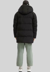 Čierna bundová vesta s kapucňou, prešívaný dizajn a dlhá dĺžka. Skombinovaná so svetlozelenými nohavicami a bielymi teniskami. Jednoduchý, funkčný štýl.