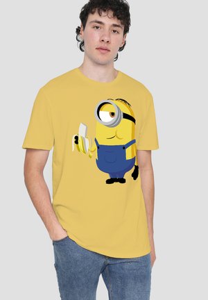 Young man wearing a yellow T-shirt with a graphic of a Minion holding a peeled banana, paired with blue jeans.