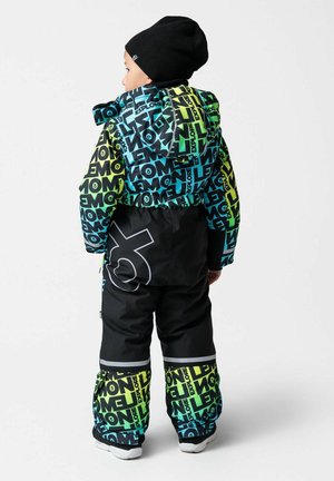 Dziecko ubrane w kolorowy, wzorzysty zimowy kombinezon z kapturem i czarną czapką, stojące tyłem na białym tle.