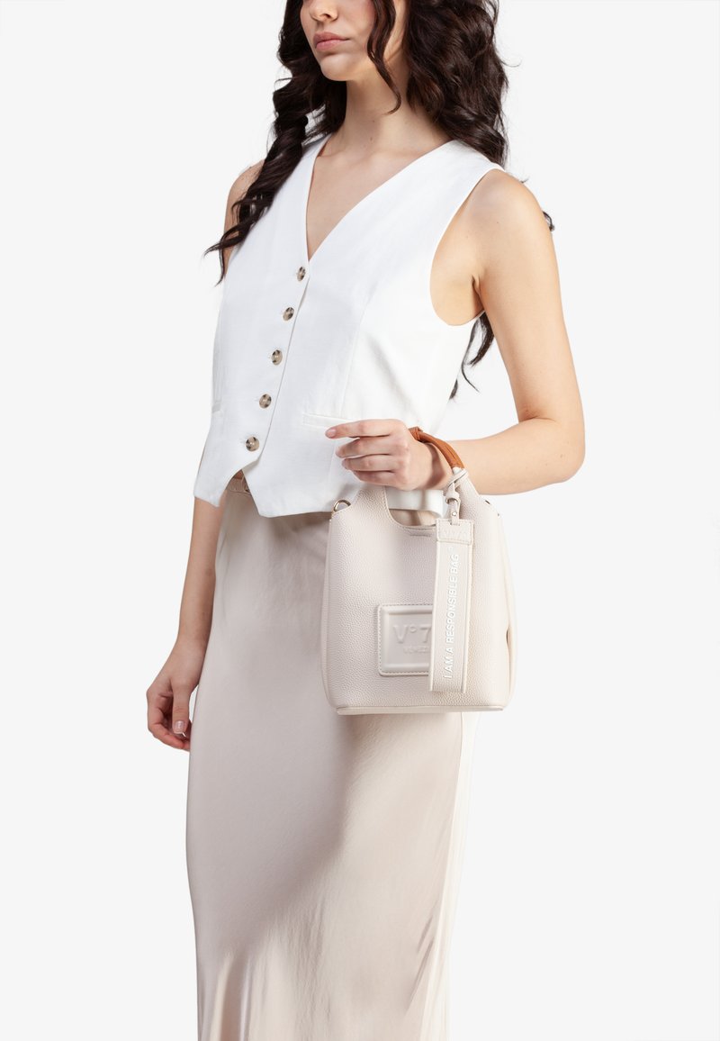 Donna in canotta bianca senza maniche e gonna beige tiene una piccola borsa color crema con manico marrone e un'etichetta che legge "Sono una borsa responsabile."