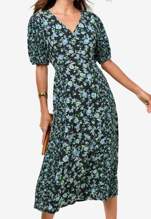 Vrouw draagt een zwarte midi-jurk met blauwe en groene bloemenprint en opgezette korte mouwen, houdt een bruine clutch in één hand.