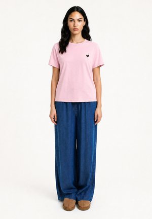Frau mit langen dunklen Haaren, die ein hellrosa T-Shirt mit einem kleinen schwarzen Herz und lockere blaue Jeans trägt, steht aufrecht.