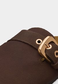 Κοντινό πλάνο ενός καφέ σουέτ παπουτσιού με φαρδιά λουράκι και λαμπερή χρυσή ορθογώνια αγκράφα.