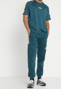 Turkos atletisk träningsdress med gula accenter; kortärmad topp och smalbyxor. Materialet är glatt och lämpligt för aktiv klädsel.