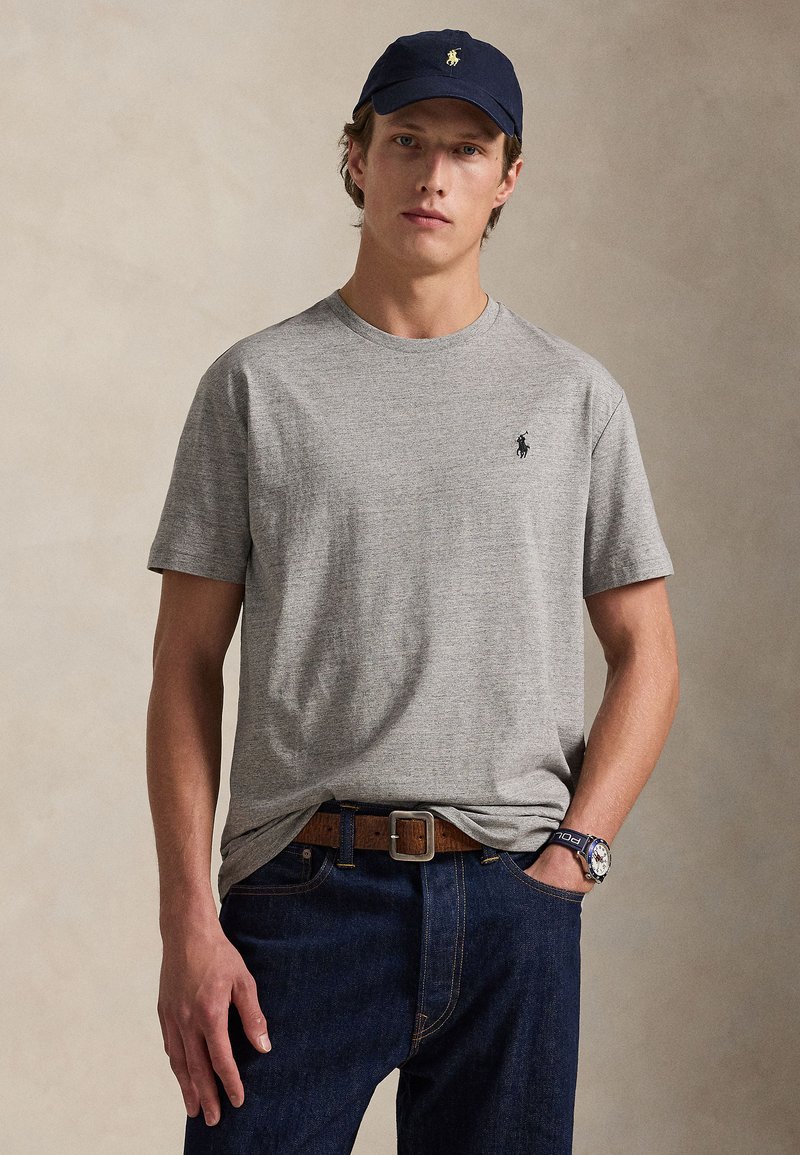 Νεαρός άνδρας που φορά γκρι μπλουζάκι Polo Ralph Lauren, ναυτικό καπέλο με λογότυπο, σκούρα μπλε τζιν, καφέ ζώνη και ρολόι χειρός, στέκεται σε ουδέτερο φόντο.