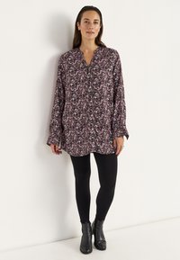 Langärmlige Blumen-Tunika in dunklem Violett mit rosa, weißen und grünen Blumen, mit einem V-Ausschnitt und Knopfleiste, kombiniert mit schwarzen Leggings.