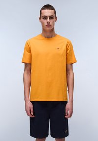 Tricou din bumbac portocalie cu guler rotund, mâneci scurte și un detaliu mic cu logo pe piept. Modelul l-a asortat cu șorturi bleumarin.