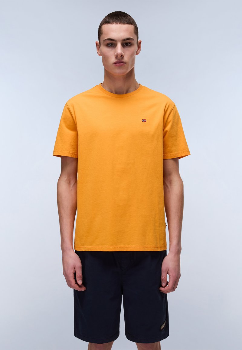 Tricou din bumbac portocalie cu guler rotund, mâneci scurte și un detaliu mic cu logo pe piept. Modelul l-a asortat cu șorturi bleumarin.