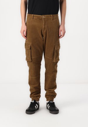 Pantalons cargo en velours côtelé marron avec une ceinture élastique, deux grandes poches latérales et une texture côtelée. Des baskets noir et blanc complètent le look.