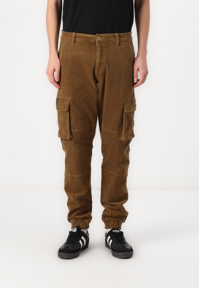 Bruine corduroy cargobroek met een elastische tailleband, twee grote zijzakken en een geribbelde textuur. Zwart-witte sneakers maken de look compleet.