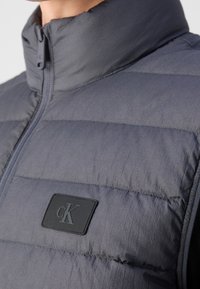 Szara kurtka puchowa z wysokim kołnierzem, z poziomą, pikowaną teksturą, z przodu zamek błyskawiczny oraz czarna gumowa naszywka z logo na piersi.
