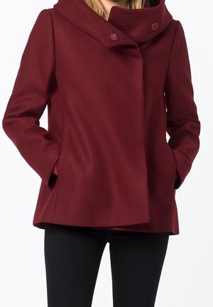 Femme portant un manteau bordeaux boutonné avec un col montant et les mains dans les poches avant, assorti à un pantalon noir.