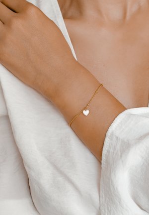 Pulsera de cadena de oro con un pequeño y brillante colgante en forma de corazón. La pulsera está delicadamente enlazada, descansando sobre un brazo ligeramente bronceado.