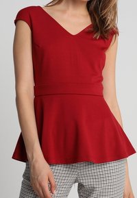 Punane peplum-top, mis on valmistatud tekstuuriga kangast, sügava V-kaelusega ja kinnastega varrukatega, koos ruuduliste pükstega.