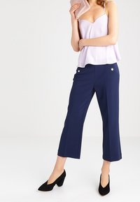 Haut sans manches lilas avec un design peplum, associé à un pantalon court bleu marine foncé avec des boutons sur le devant, et des escarpins noirs à bout pointu.