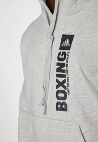 Felpa grigia con cappuccio, dotata di cordoncino, tasca frontale e testo stampato in nero audace: "BOXING" e "ONLY THE BEST FOR THE ATHLETE", con logo Adidas.
