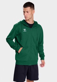 Groene hoodie met rits gemaakt van zachte stof, met een zwart logo op de bovenste linkerkant van de borst, kangoeroezakken en een verstelbare trekkoordkap.
