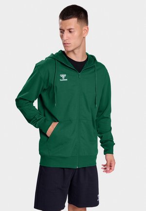 Haut de sweatshirt zippé vert en tissu doux, arborant un logo noir sur la poitrine gauche, des poches kangourou et une capuche avec cordon de serrage ajustable.