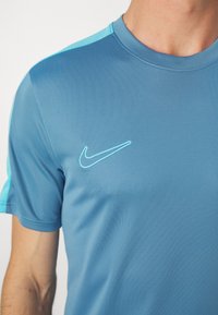 Modré sportovní tričko s krátkým rukávem, které má na rameni světle modrý akcent a na hrudi vyšívané logo Nike ve světle modré barvě.