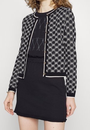 Femme portant un cardigan noir à boutons à motifs par-dessus une robe noire à encolure ronde et des boucles d'oreilles dorées.